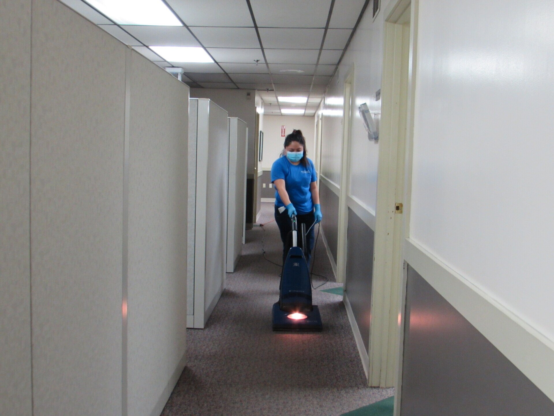 Woman in mask vacuums office hallway. commercial cleaning in progress.